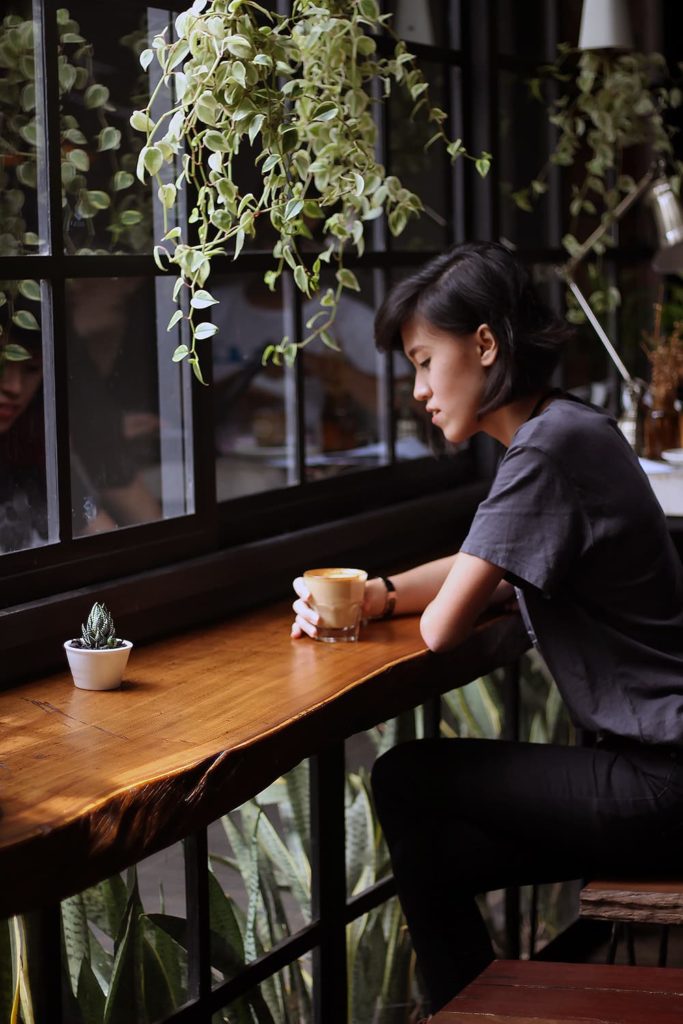 Image of a woman sitting at a bar top with a plant hanging over her head and holding a cup of coffee. When working with an anxiety therapist in Los Angeles, CA, you can learn to cope with loneliness. | 91006 | 90071