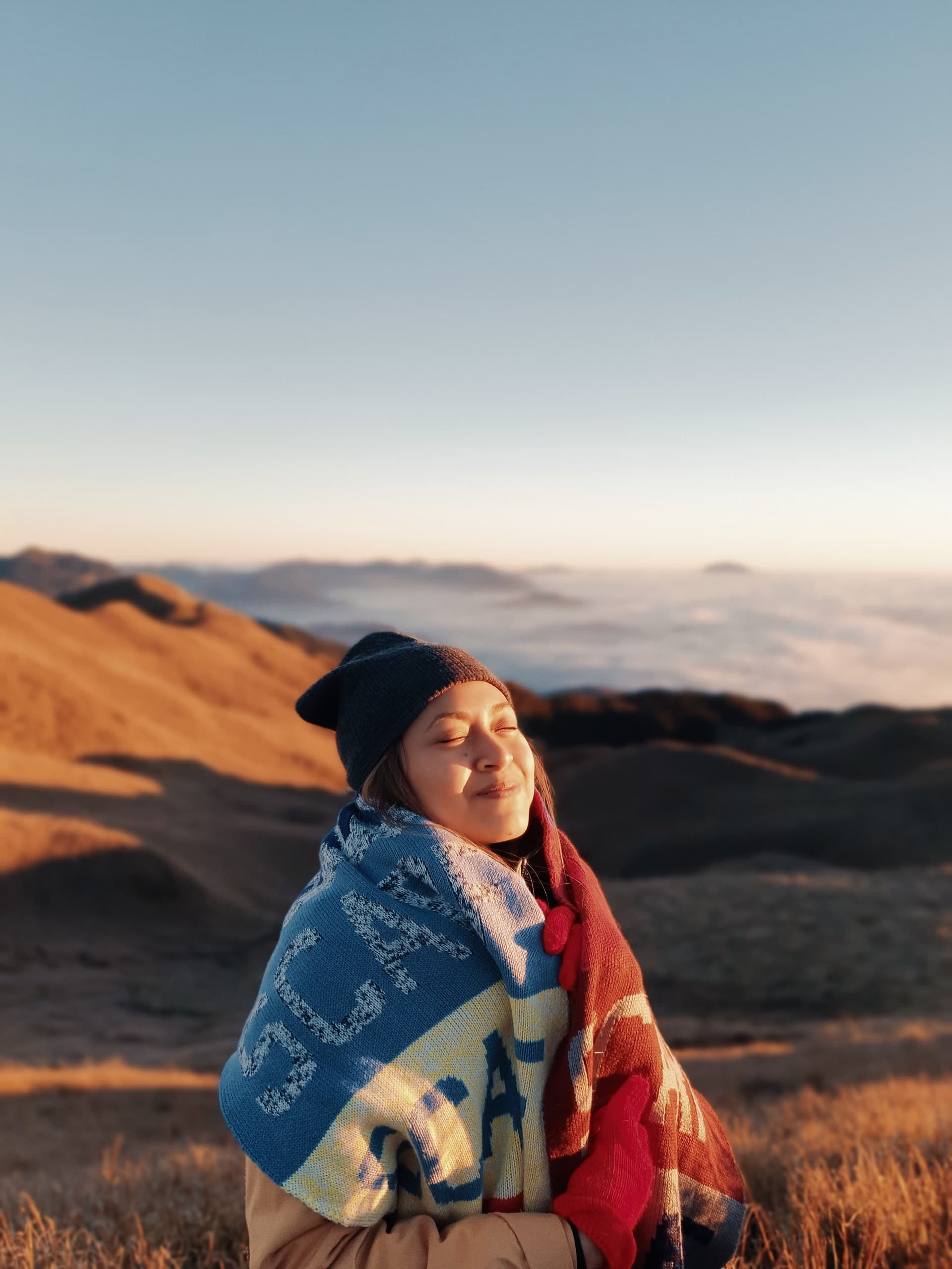 Photo of a young woman snuggling in a blanket outdoors and smiling. Those experiencing symptoms of trauma can benefit from working with a therapist for trauma therapy and PTSD treatment in Los Angeles, CA. 90071 | 90232