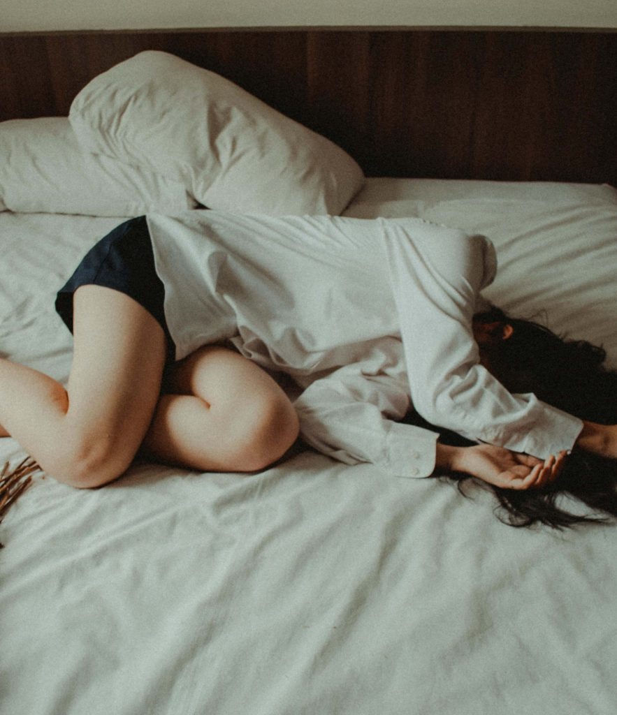 Image of a woman lying in bed with her arms covering her face. This woman represents the effects of perinatal mood disorders on mothers. Postpartum therapy in Los Angeles, CA can help mothers overcome perinatal mood disorders. | 91006 | 90071