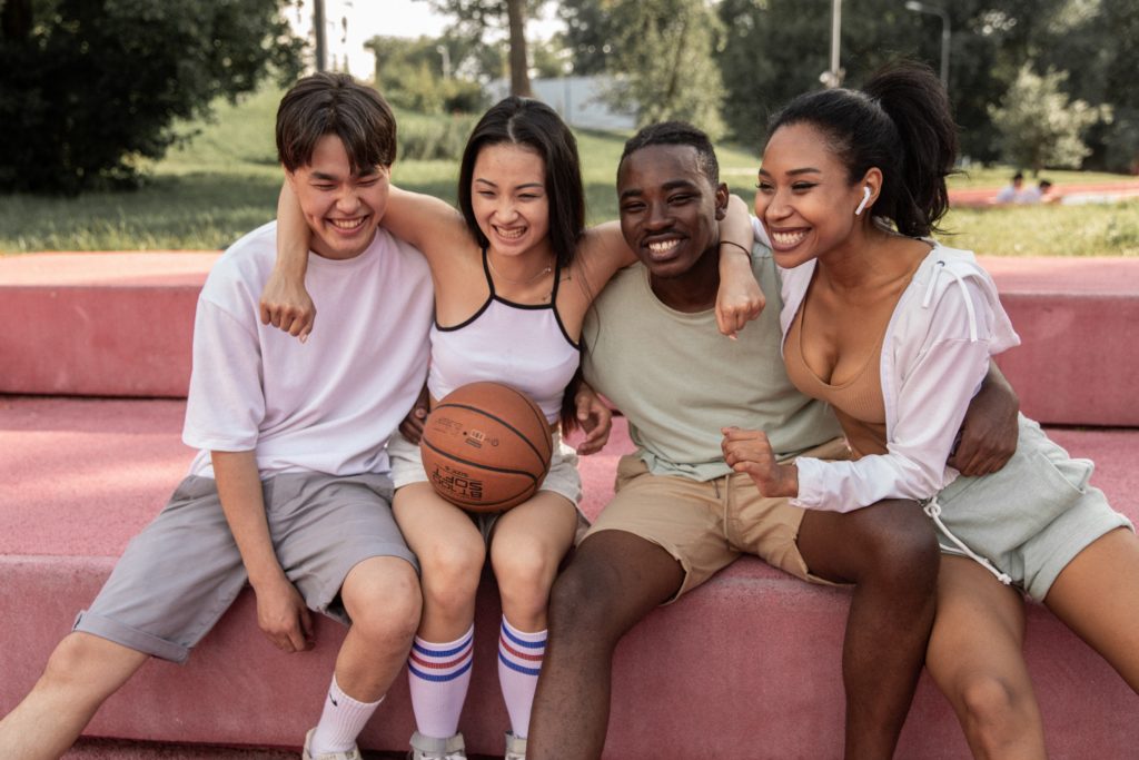 Happy multiracial teenagers with their friends embracing on bench after basketball training representing how Yellow Chair Collective can provide excellent Asian American teen therapy for Asian and Asian American teens in Southern California.