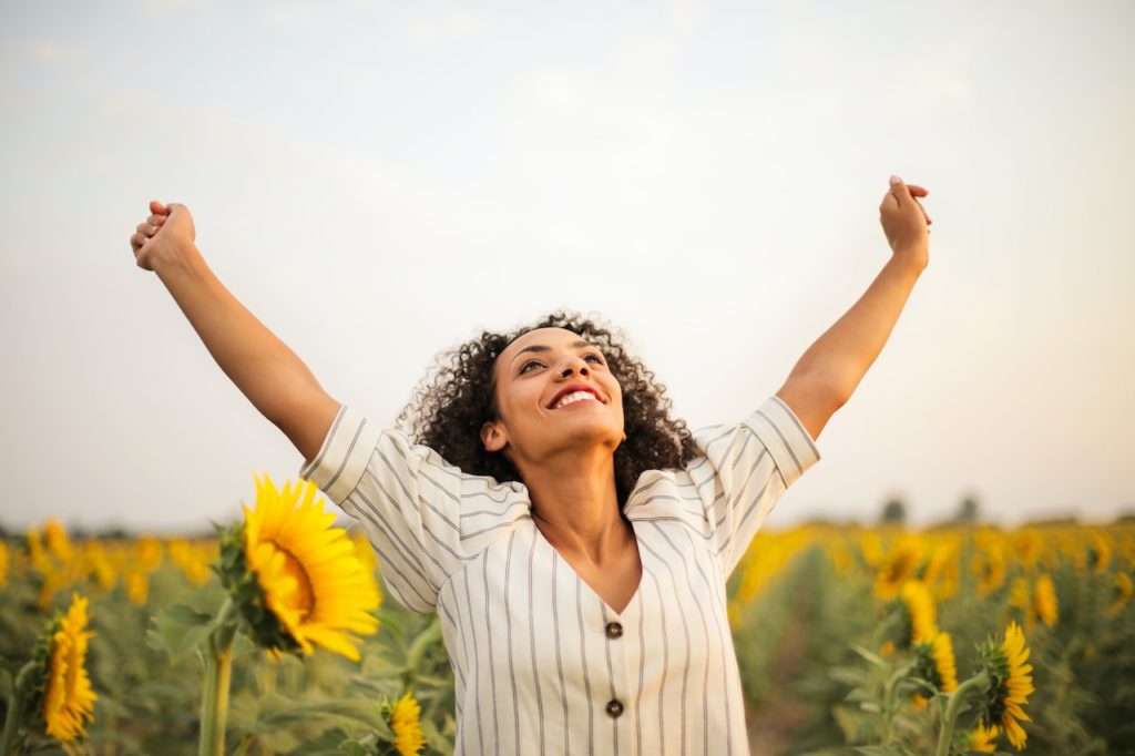 A Black woman stretching her arms in a sunflower field representing the freedom she feels after receiving therapy and counseling sessions from an anxiety therapist in New York City.