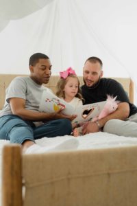 A multicultural, gay couple reading to their daughter on a couch representing that understanding cultural differences with your partner can lead to better family dyanamics.