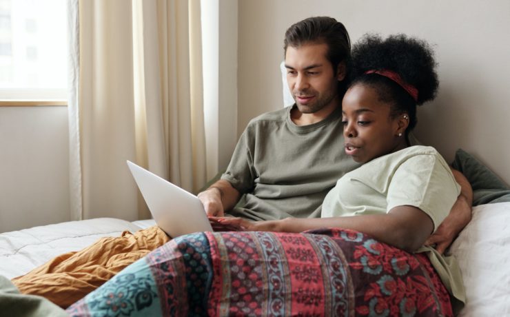 Multicultural couple reading off of a laptop in bed representing how understanding their cultural differences can be an opportunity to grow closer in their relationship.