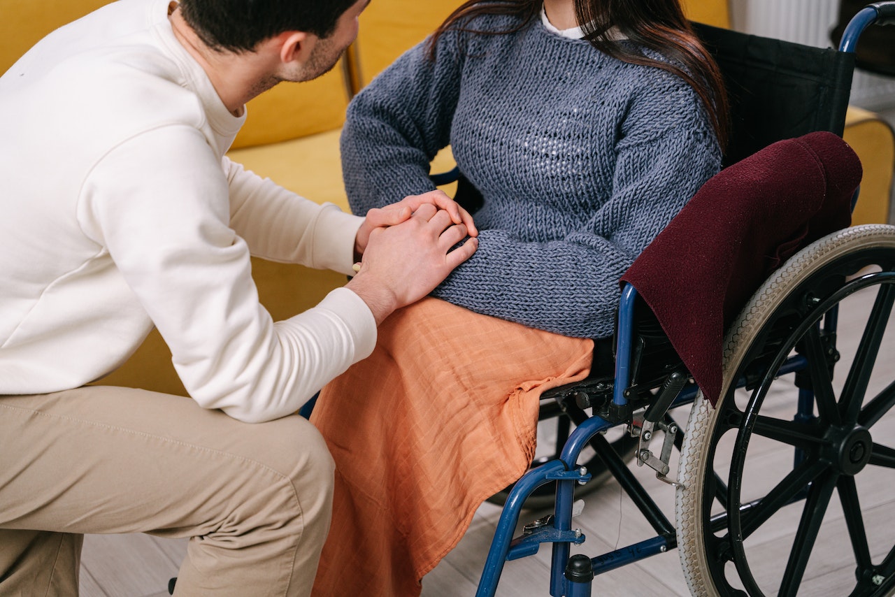 A man who is a caregiver holding the hand of a woman who is using a wheelchair representing the social support that caregivers need and can find from therapy for caregivers in New York.