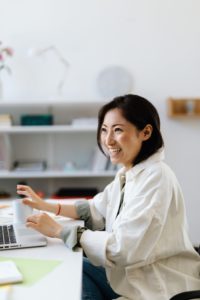 A woman smiles while sitting at a laptop workspace. This could symbolize the newfound joy cultivated by adult ADHD therapy in Los Angeles, CA. Learn more about therapy for ADHD in Los Angeles, CA, and other services offered in person and online via online therapy in California. 
