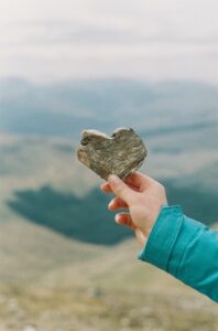 A person holds up a rock in the shape of a heart in the mountains. This could represent the self-love cultivated by working with an Asian American therapist in Los Angeles. Search for Asian American therapy near me today. 
