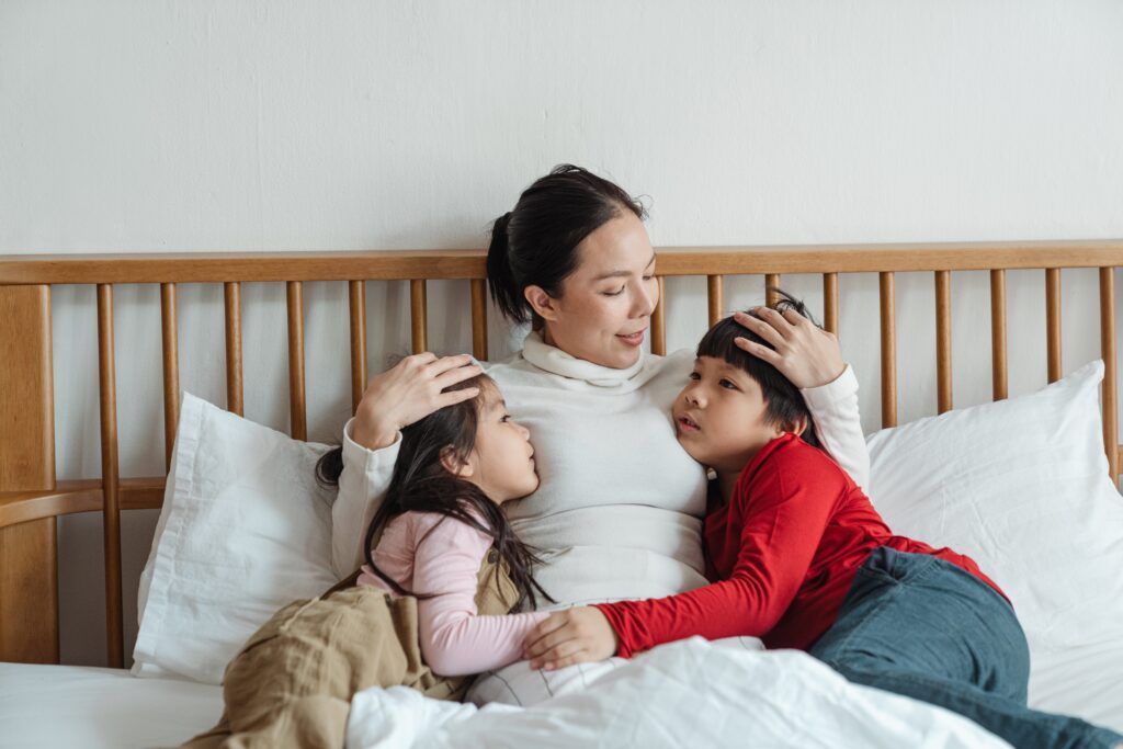 An Asian woman sitting in bed, holding two young children. Learn how an Asian American therapist in Los Angeles, CA can offer therapy for parenthood in Los Angeles, CA and other services. Search for Asian American pregnancy and postpartum therapy to learn more.