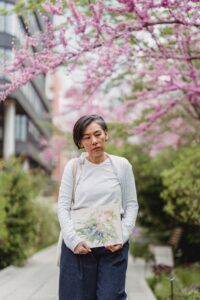 An Asian American women carries a painting while walking under a cherry blossom tree. Learn how an Asian American therapist can help you address anxiety via anxiety treatment in Los Angeles, CA. Search for online anxiety treatment in California today.
