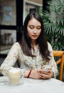 An Asian American woman sits at a table while looking at her phone. This could represent the search for an Asian American therapist in Los Angeles. Search for therapy for Asian Women in Los Angeles, CA or Asian American therapy near me today.
