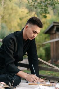 A man focuses on painting while sitting outside in a forest. This could represent the benefits of art therapy when working with an Asian American therapist. Learn more about the help an Asian American therapist in California can offer by searching for Asian American therapy today.
