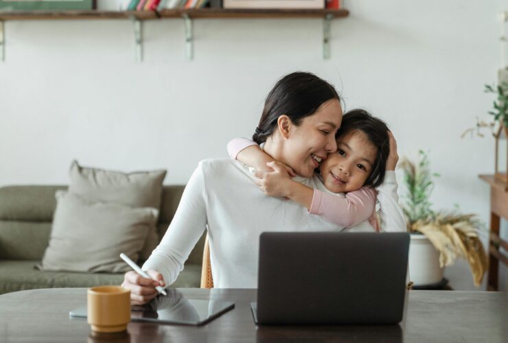 A mother sitting at her laptop table smiles while her daughter hugs her. This could represent the benefits of working with an Asian American therapist in California. Learn more about how therapy for asian women in Los Angeles, CA can offer support by searching for individual therapy in Los Angeles, CA.