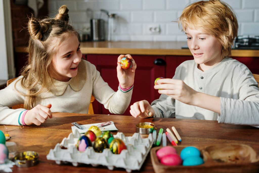 Two children, a girl and a boy, sit at a table with various art supplies, decorating Easter eggs. They smile while holding and admiring their painted eggs in a bright kitchen setting. Learn how a neurodiversity-affirming therapist in los angeles, ca can offer support by contacting an Asian American therapist today. Search for neurodiverse affirming therapy los angeles, ca today.