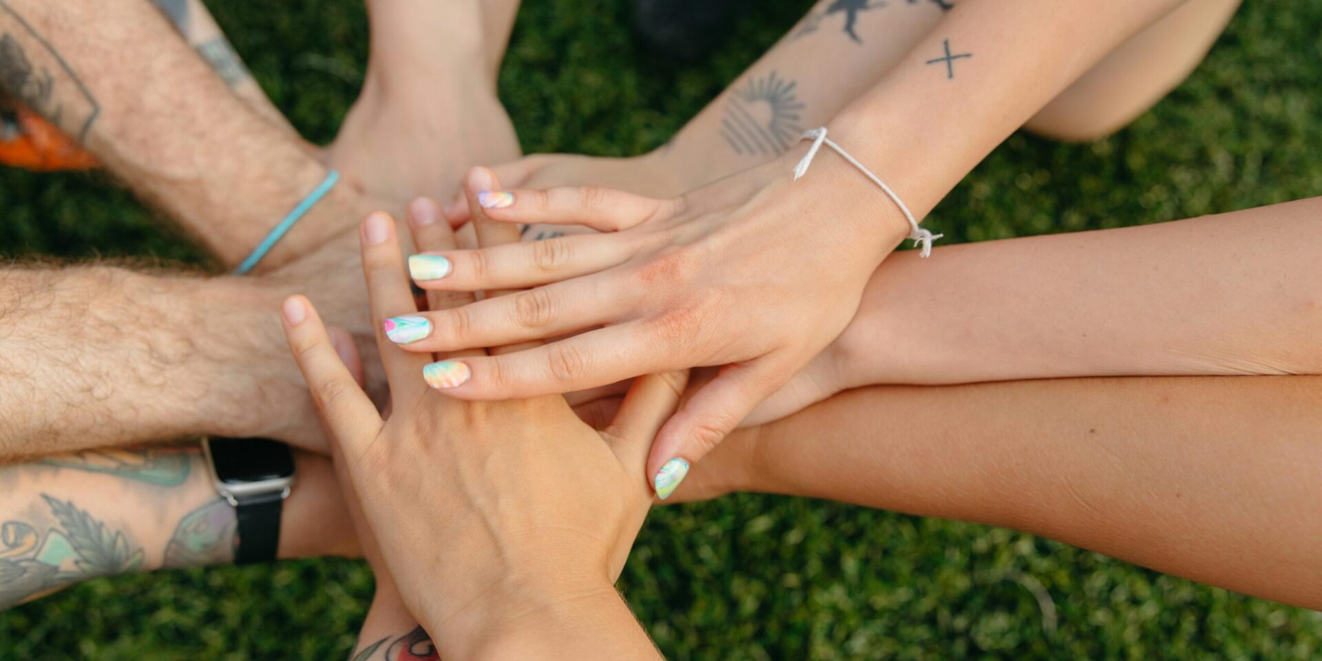 An image of many hands with different skin tones and apperances, overlapping one another over a grassy area. An image that illustrates the way our many different identities intersect.