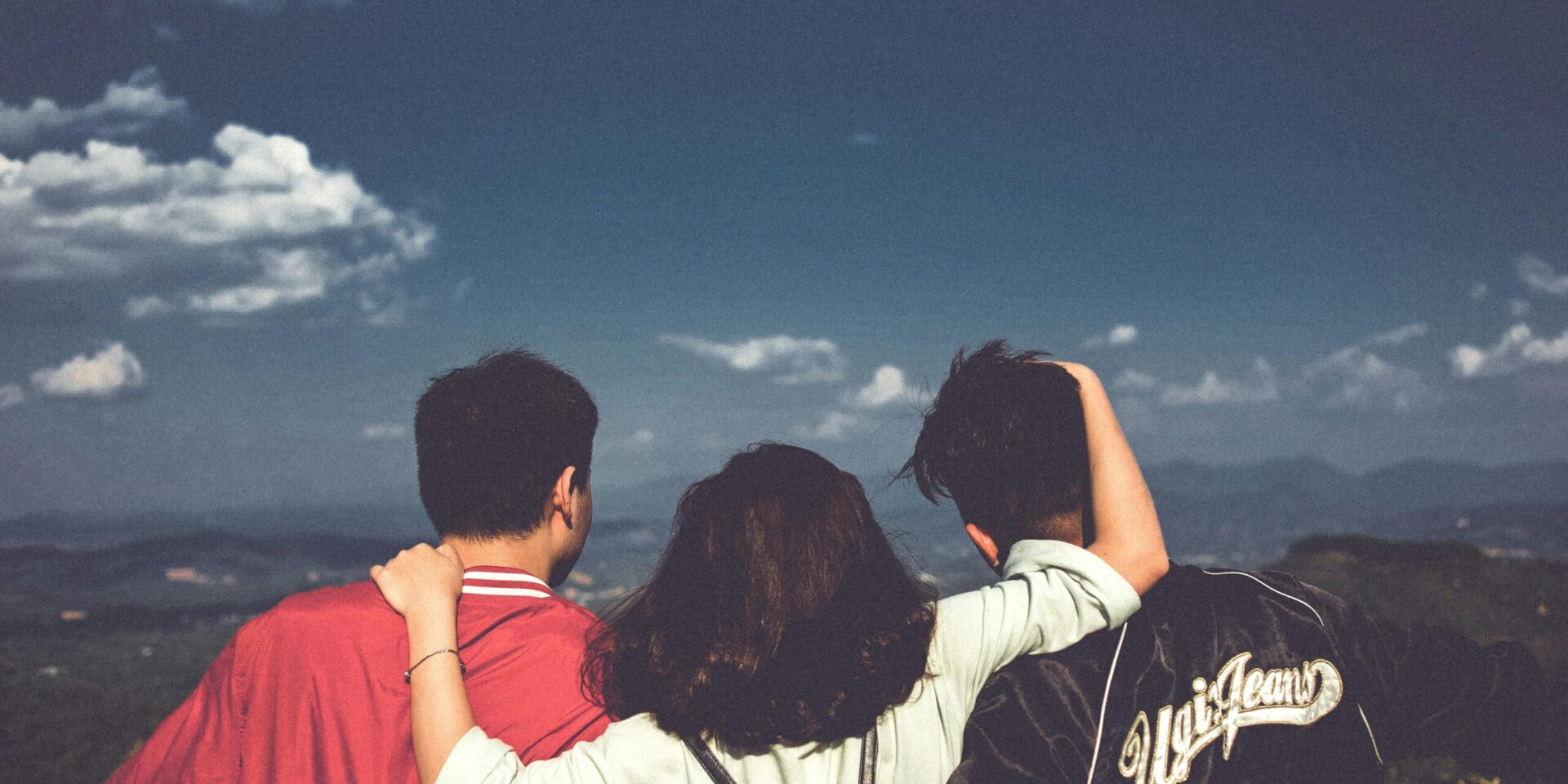 Three people standing together with their backs to the viewer, two men on either side, one woman in the middle with her arms around them, as they look out at a blue sky and a valley.