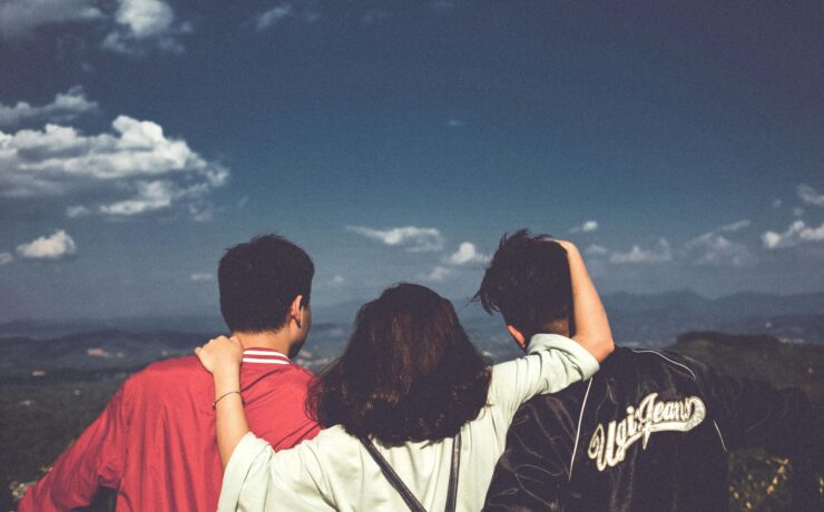 Three people standing together with their backs to the viewer, two men on either side, one woman in the middle with her arms around them, as they look out at a blue sky and a valley.