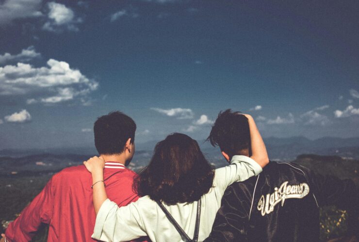 Three people standing together with their backs to the viewer, two men on either side, one woman in the middle with her arms around them, as they look out at a blue sky and a valley.