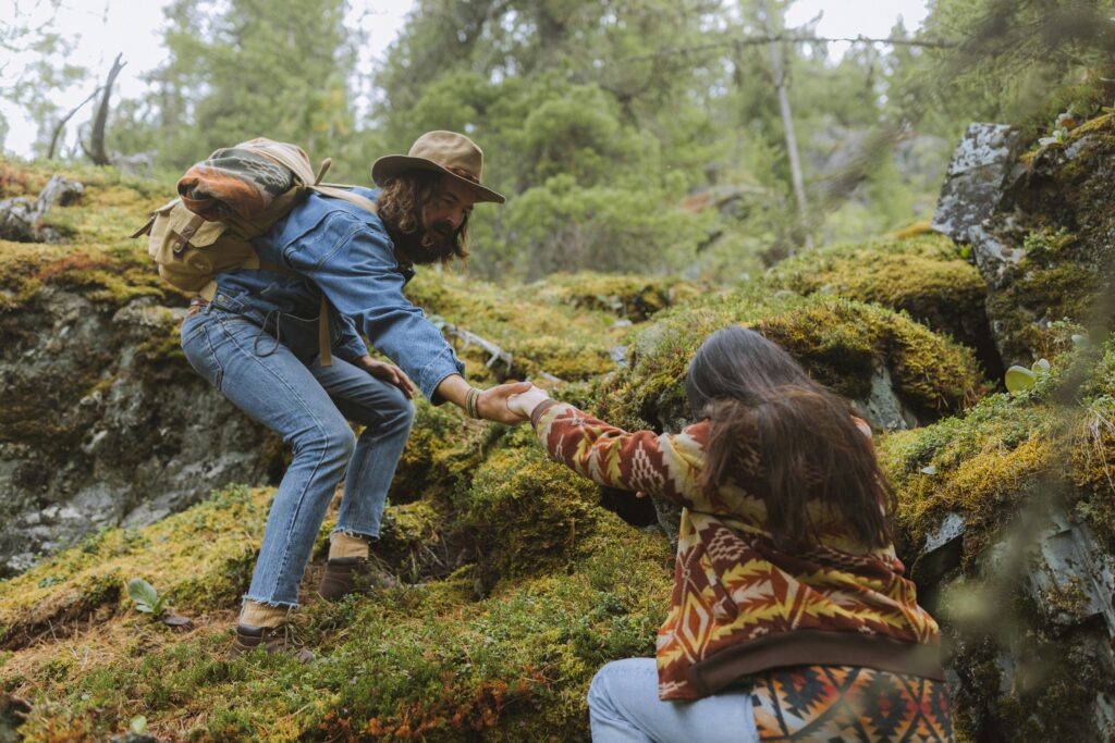 A couple trying to climb a grassy hill, the man above extending a hand to the woman below to help her up the hill.