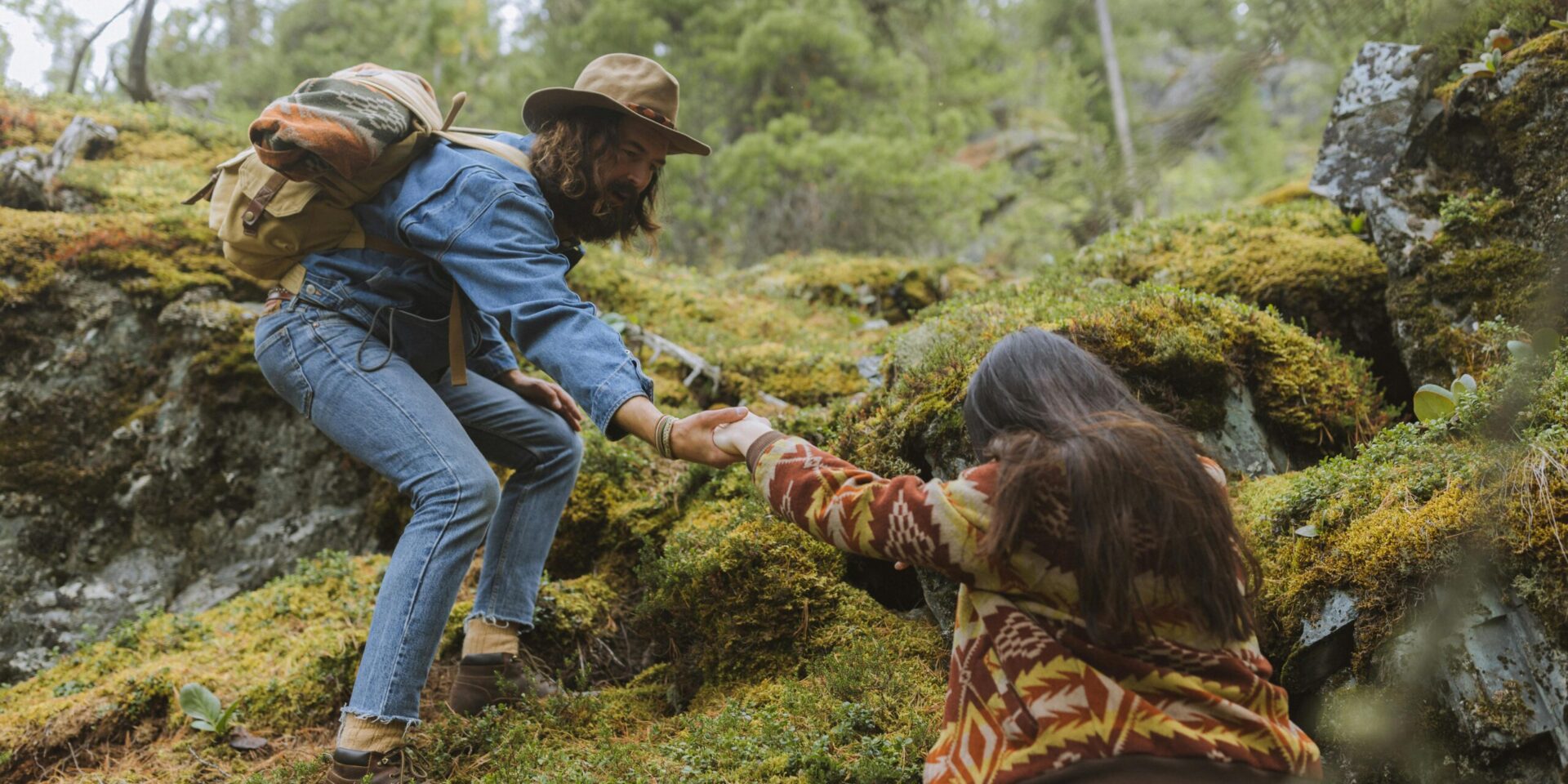 A couple trying to climb a grassy hill, the man above extending a hand to the woman below to help her up the hill.