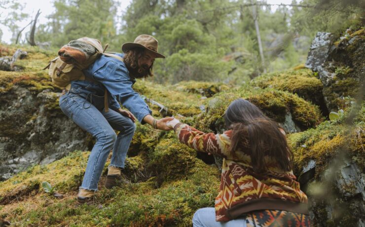 A couple trying to climb a grassy hill, the man above extending a hand to the woman below to help her up the hill.