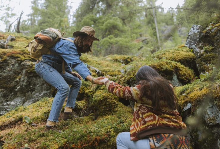 A couple trying to climb a grassy hill, the man above extending a hand to the woman below to help her up the hill.