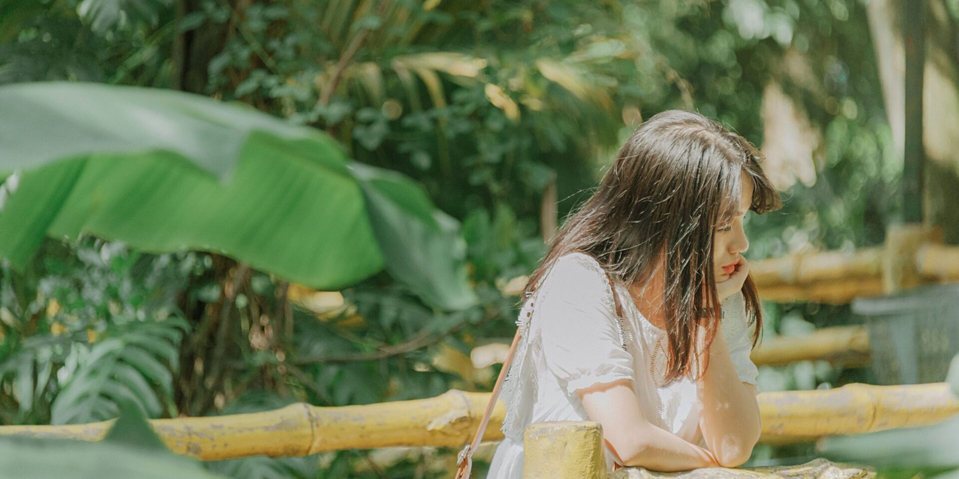 A young Asian woman leans on a bamboo railing by herself with her chin resting on her hand. She looks pensive.