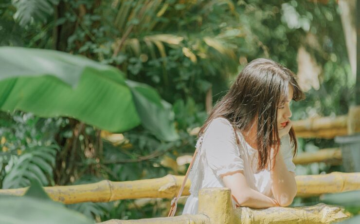 A young Asian woman leans on a bamboo railing by herself with her chin resting on her hand. She looks pensive.