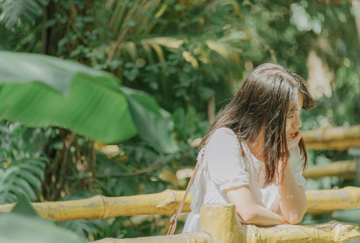 A young Asian woman leans on a bamboo railing by herself with her chin resting on her hand. She looks pensive.