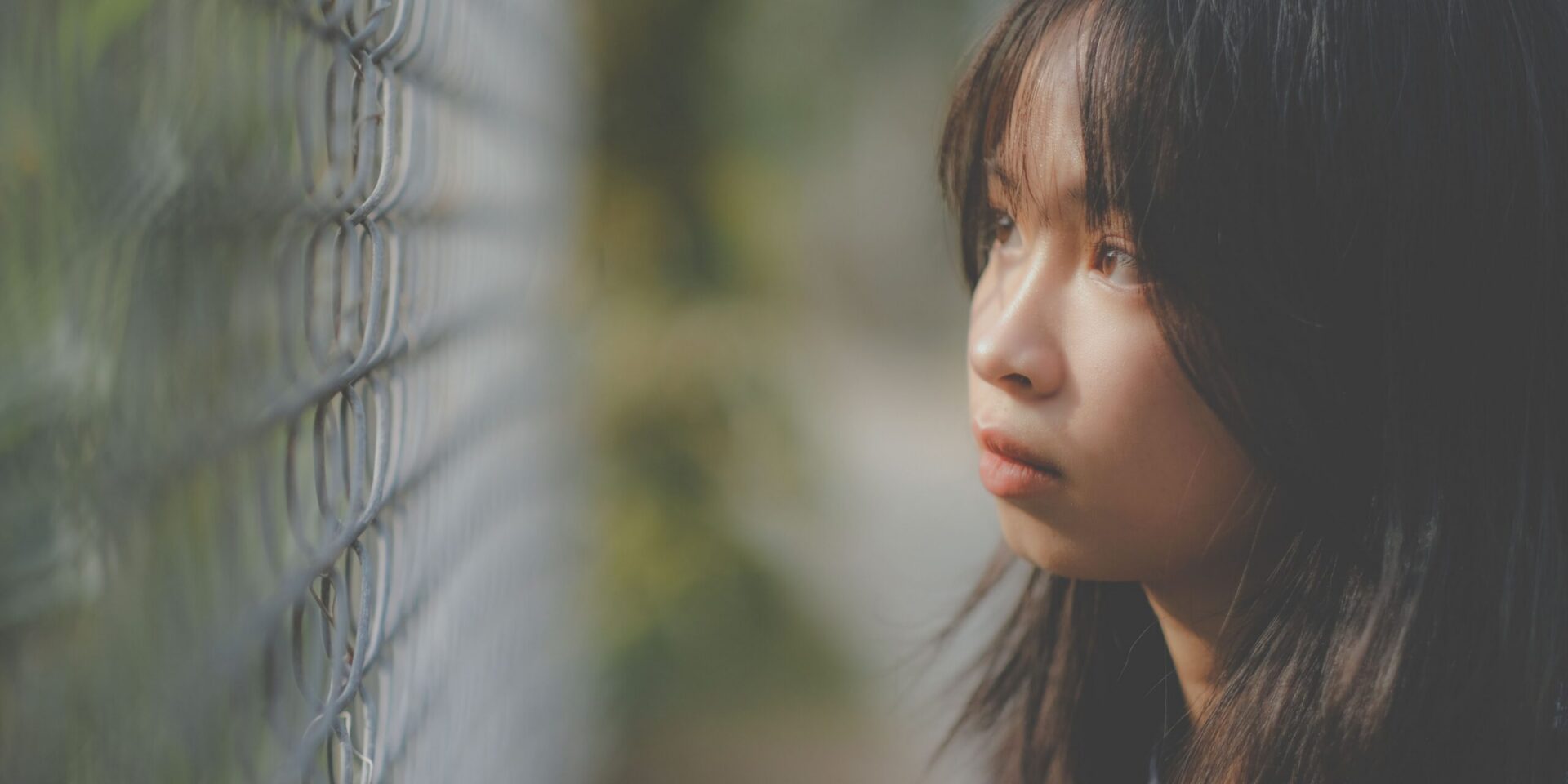 A young Asian girl looks through a wired fence looking lost in thought.