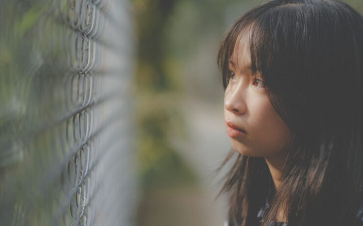 A young Asian girl looks through a wired fence looking lost in thought.