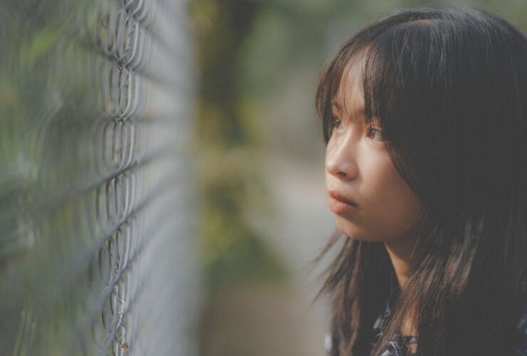 A young Asian girl looks through a wired fence looking lost in thought.