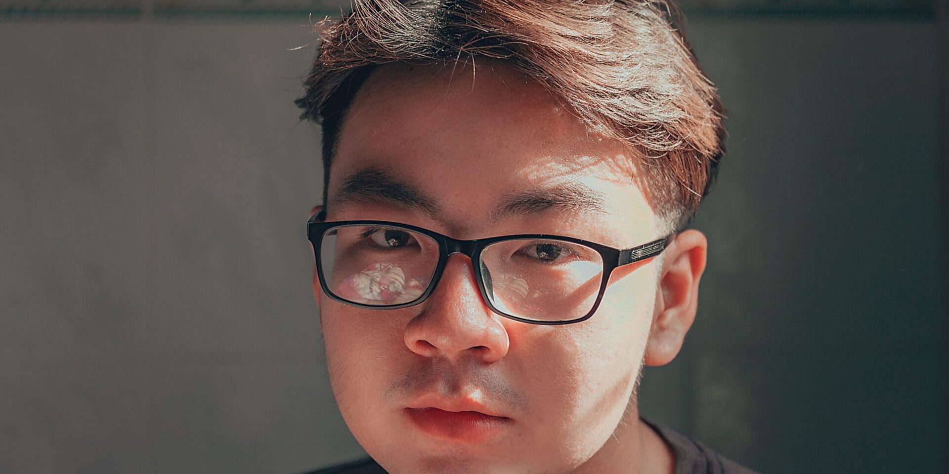 A young Asian man in thick black rimmed glasses looks seriously at the camera.