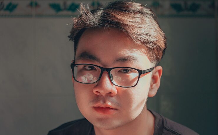 A young Asian man in thick black rimmed glasses looks seriously at the camera.