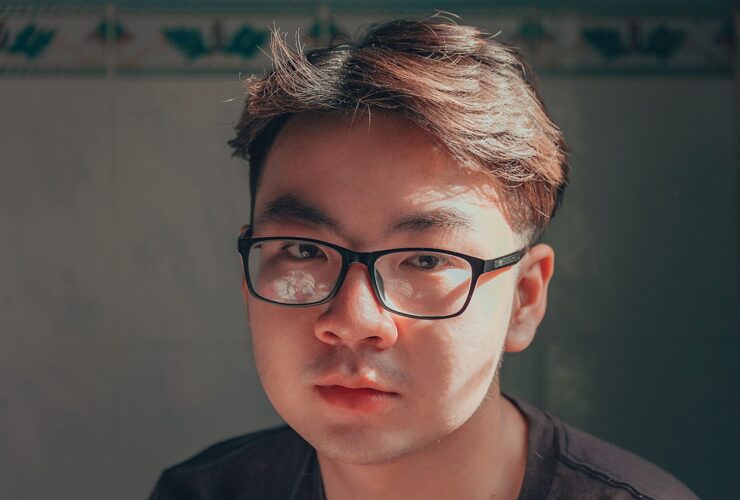 A young Asian man in thick black rimmed glasses looks seriously at the camera.