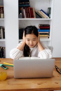 pexels-shkrabaanthony-6267055 A young Asian girl sits in front of her laptop with her hands covering her ears. She looks stressed.