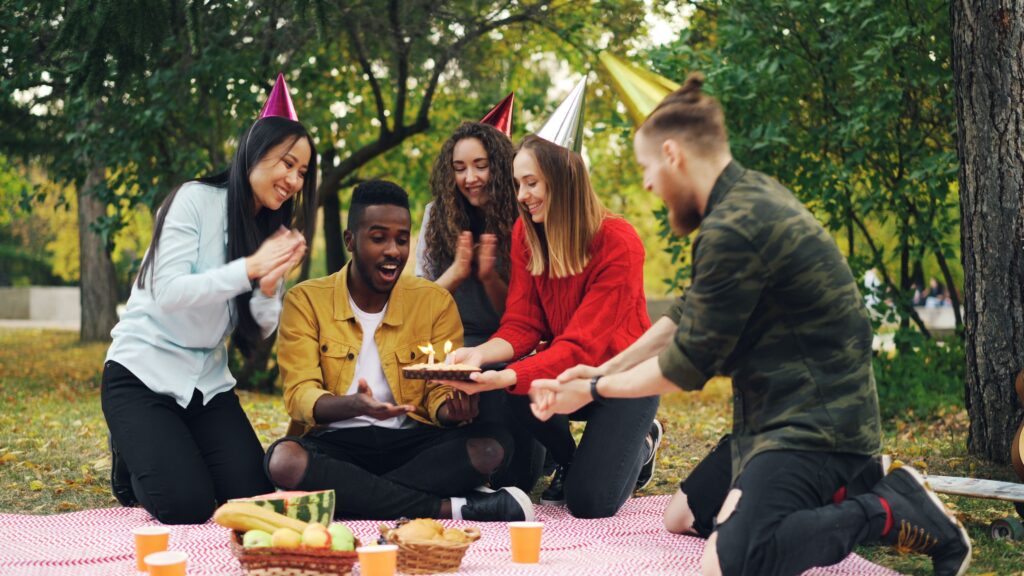 An ethnically diverse group of friends (seeming 20s-30s in age) are having a picnic birthday party.