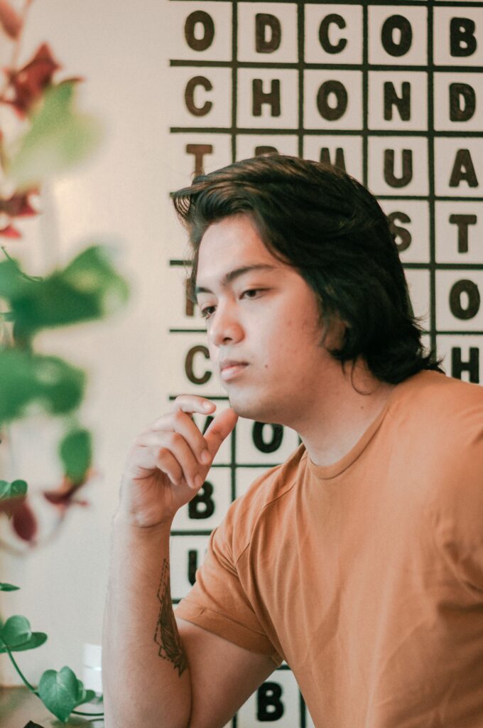 pexels-thegiansepillo-1595943-6772436 A young Asian man in an orange shirt sits looking thoughtful at what seems to be a restaurant or cafe.